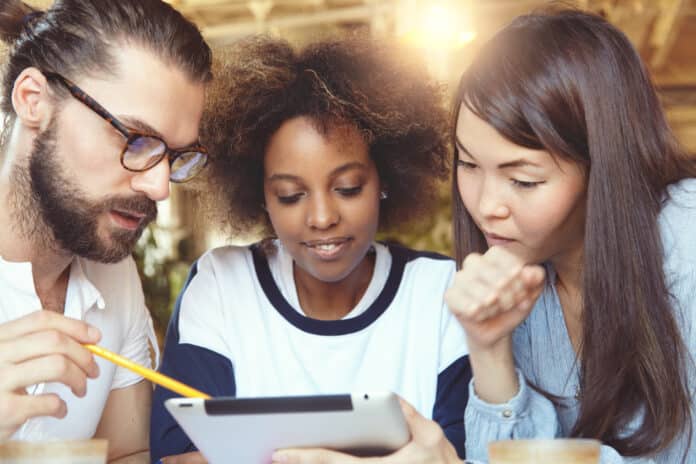International team. African and Asian females at cafe listening with attention to their male partner in glasses, presenting business strategy on tablet, showing information on screen with pen
